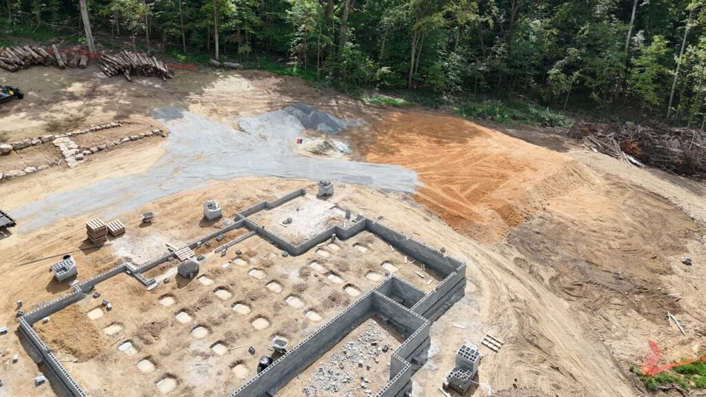 Aerial view of a new brown house and attached red barn situated on a plot of recently tilled brown earth, bordered by green fields, with a black dump truck driving on the paved road and rolling hills in the background under a blue sky.
