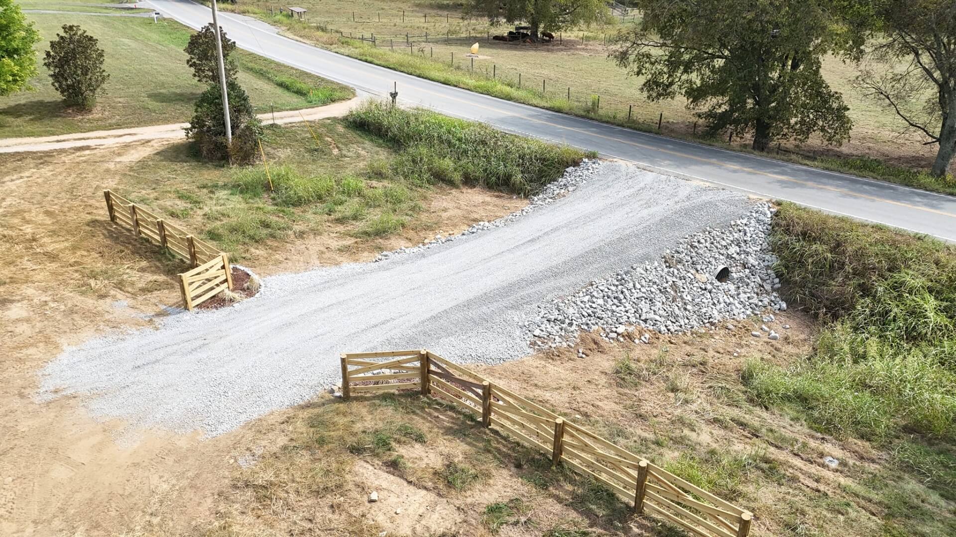 Ground-level view of a new construction site showing a large pile of gray crushed stone in the foreground, bordering a cleared area of reddish-brown dirt, set against a dense backdrop of lush green trees under an overcast sky.