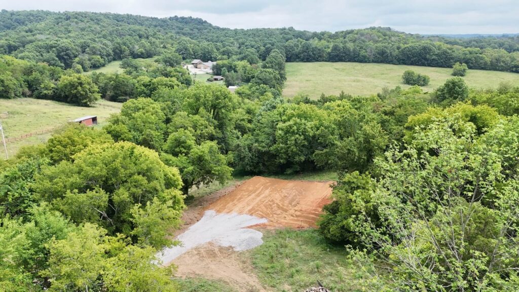 View from a piece of heavy equipment showing the raised black steel arm in the foreground, over a field of dry, brown grass and dirt, with a dense line of winter trees with bare branches under a hazy, late-day sky.