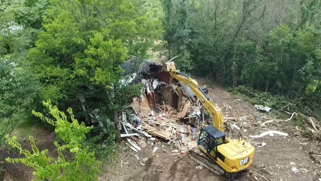 Aerial view of a construction clearing on a hillside, showing a patch of reddish-brown dirt and a pile of gray crushed stone, surrounded by lush green deciduous trees with a farmhouse visible in the distance.