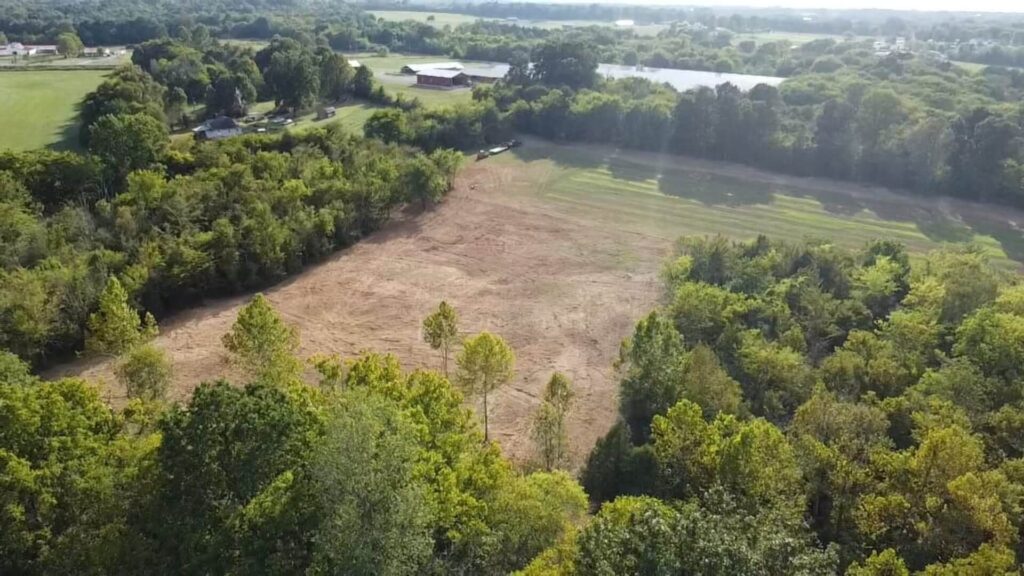View from a piece of heavy equipment showing the black steel boom and bucket attachment over a field of dry, brown grass and dirt, with a line of bare trees in the distance under a hazy, late afternoon or winter sky.