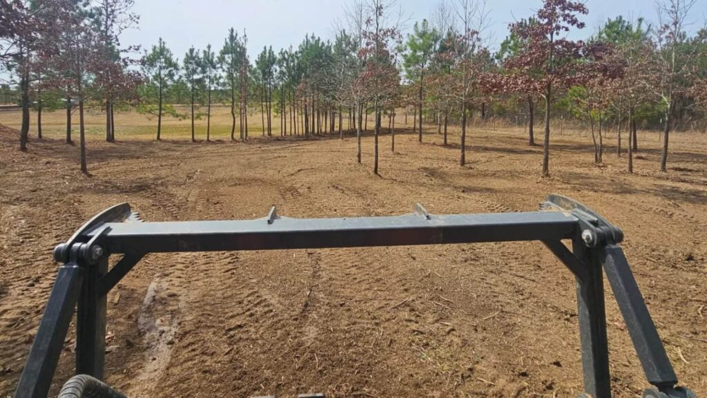 Aerial view of a new brown house and attached red barn on a plot of recently tilled brown earth, bordered by green fields, with a dump truck driving along the road in the foreground and rolling hills in the distance.