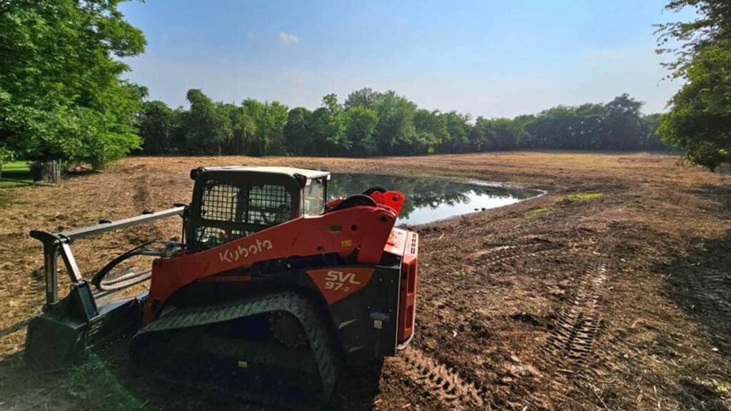 Wide-angle, low perspective photograph of a large, flat brown field covered in crop residue, framed by a dense line of summer trees under a brilliant blue sky with expansive white clouds.