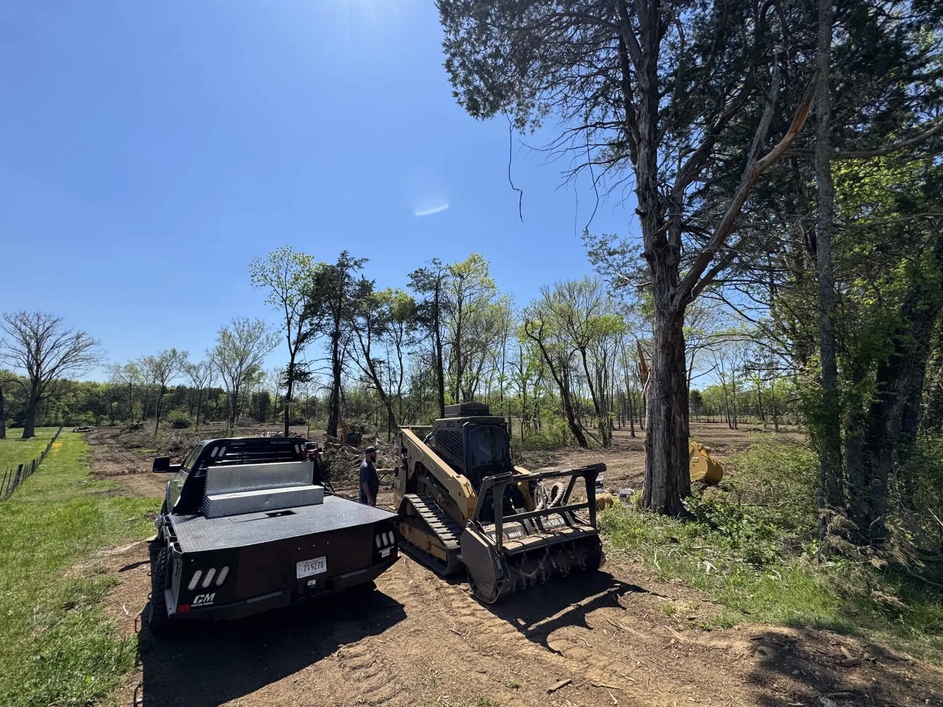 A black flatbed work truck with a silver toolbox is parked on a dirt path next to a yellow Caterpillar skid steer equipped with a forestry mulching head. A man stands between the two vehicles. The scene is set on a sunny day in a rural area with tall trees, green grass, and cleared land in the background under a clear blue sky.